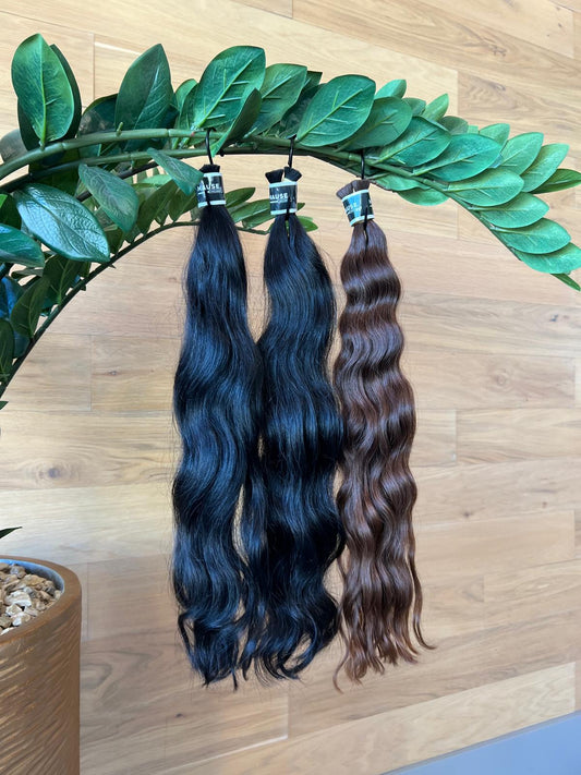 Two natural wave bulk hair extensions, one blue and one brown, hanging against a wooden background with green leaves.